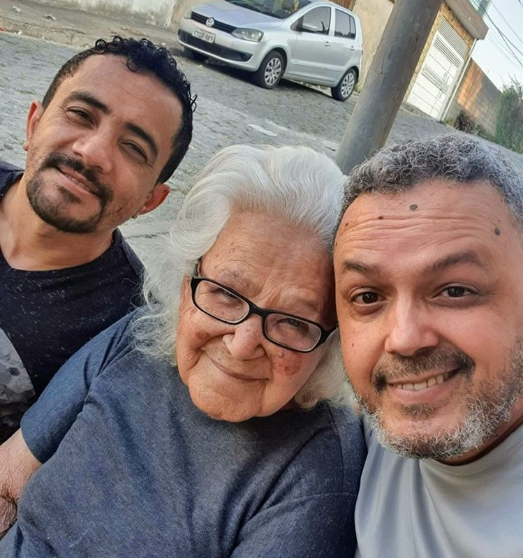David está ao lado de uma senhora e um homem. Todos estão sorrindo. Ao fundo há um carro cinza parado.